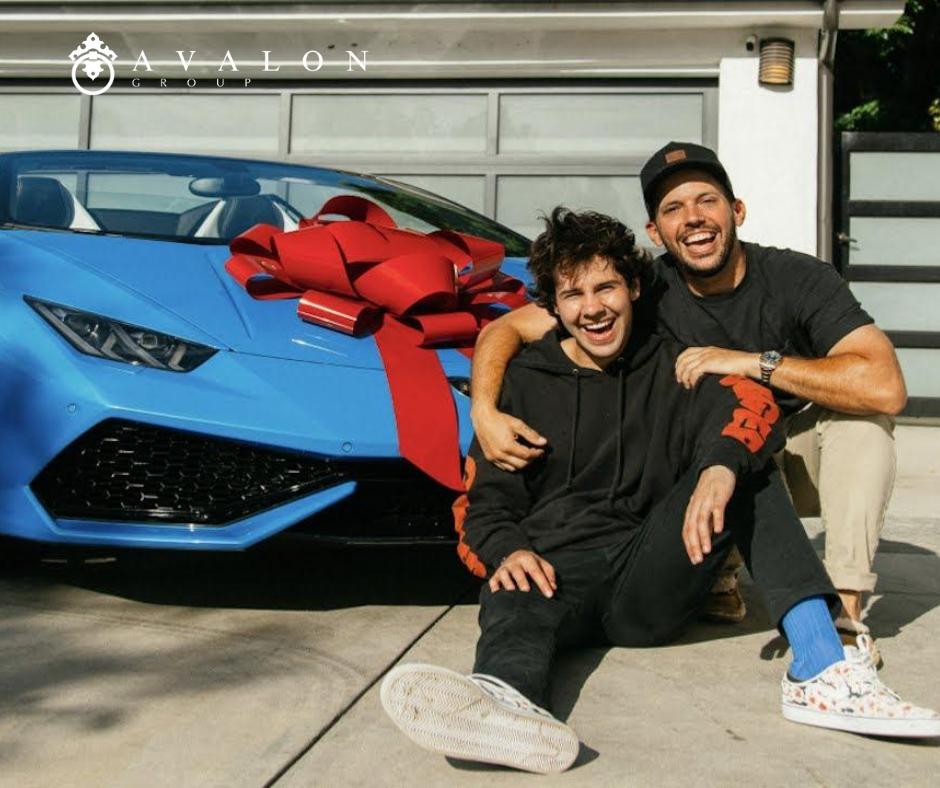 And there is a blue luxury sports car in the driveway. A gay couple is posing in front of their new car. Additionally there is a red bow on the car.