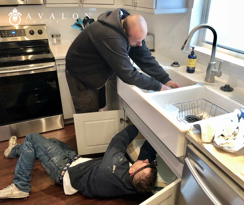 Additionally, two plumbers are working on a kitchen sink that is white. And one is under the sink and one is above. Also, the plumbers are wearing navy hoodies and blue jeans.