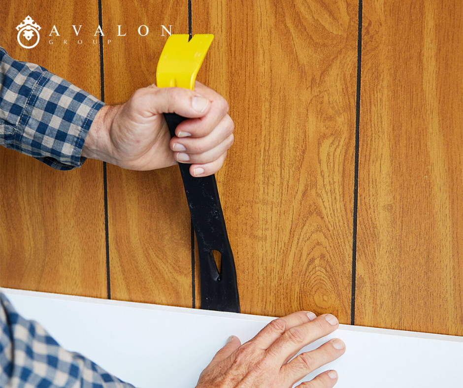 And a man is removing the baseboard in preparation of removing panelling. The man is wearing a blue plain shirt.