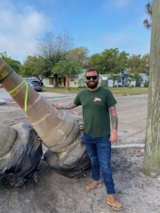 Also, Bryan Shows the Big Base of a Royal Palm He is About to Install. He has a full beard and wearing a green shirt and blue jeans.