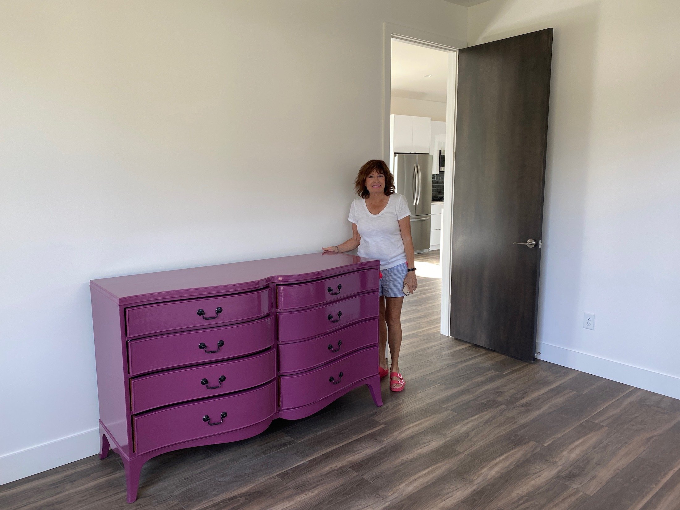Moreover, the new mulberry colored dresser creates a happy room. Aunt Pam is standing beside it in the picture. Also, There are white walls and a black door.