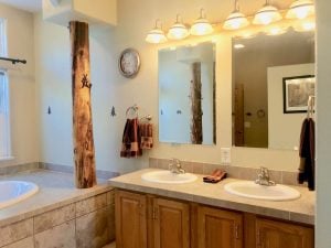 Bathroom vanity with double sink and mirrors, oval drop-in tub with wood pillar to the left