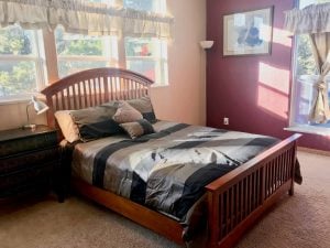 Full-size bed with headboard in a carpeted room with one wall painted burgundy and the other white, big windows letting in lots of light