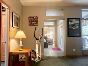 Room with dark green carpet, small table with lamp, stair-stepper exercise machine in the corner. There are 3 big pictures hanging on the wall, and you can see through the length of the house