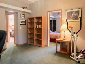 Room with dark green carpet showing open doors to 2 bedrooms; there's a small table with lamp, a stair-stepper exercise machine, and a mostly empty bookshelf